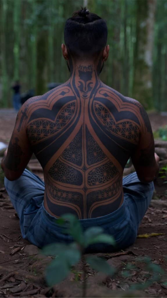 Octavio seated in the forest, back to camera, showing detailed geometric tattoo work.